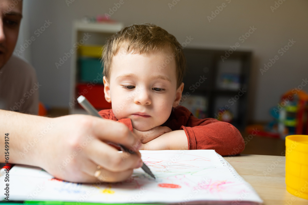A cute little toddler boy of two years old with dad draws with markers in the album in the children's room at home. Spending time with children. Educational activities for kids