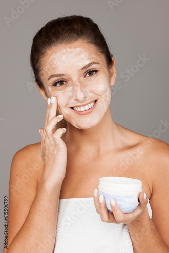 Content woman using nourishing cream from pot on hand and looking at camera isolated on gray background.