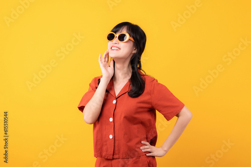 Portrait young beautiful asian woman happy smile dressed in orange clothes and sunglasses looking celebrate with hand on her face isolated on yellow studio background. © Jirawatfoto