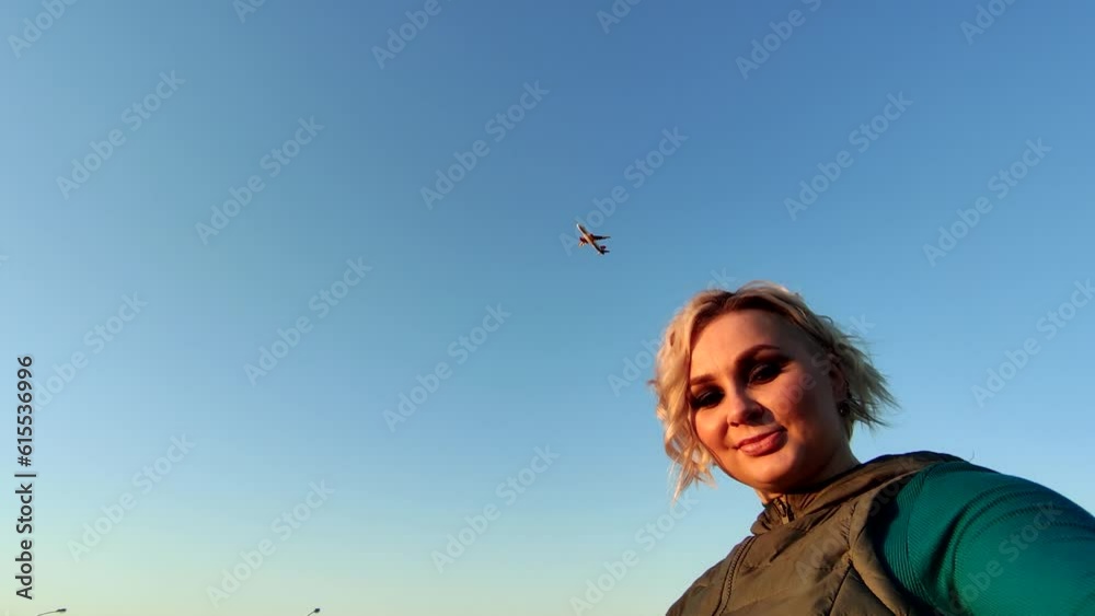 Beautiful plus-size blond woman takes a selfie video with airplane departure in sky by airfield on smartphone at sunset. Clear blue sky. Slow motion video. People and transport theme. Beautiful plus-size blond woman takes a selfie video with airplane departure in sky by airfield on smartphone at sunset. Clear blue sky. Slow motion video. People and transport theme.