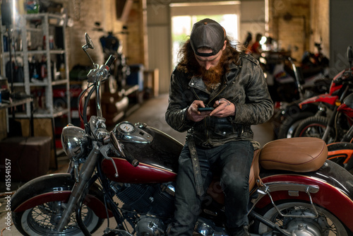 Creative authentic motorcycle workshop Garage brutal serious bearded redhead biker mechanic sitting on motorcycle with phone in hands © Guys Who Shoot