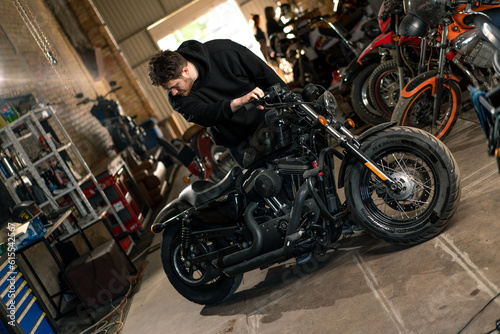 serious bearded biker in black hoodie in motorcycle workshop standing near motorcycle inspecting it for repair