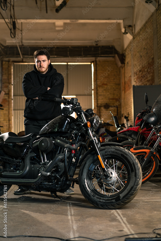 serious bearded biker in black hoodie in real motorcycle workshop garage standing near motorcycle with crossed arms