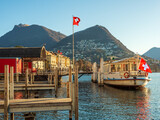 lakefront in Lugano Switzerland, with a ferry anchored at the dock, Swiss flags in the wind, and mountains in the background, in the early morning light