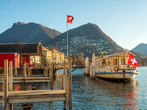 lakefront in Lugano Switzerland, with a ferry anchored at the dock, Swiss flags in the wind, and mountains in the background, in the early morning light photo