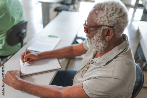 homem idoso Brasileiro negro de cabelos brancos fazendo anotações no caderno durante aula em um curso para adultos no Brasil