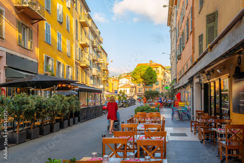 Fototapeta Naklejka Na Ścianę i Meble -  A young woman pushes a baby stroller down a narrow street of picturesque shops and sidewalk cafes in the colorful old town View Nice district of Nice, France, along the Cote d'Azur French Riviera.