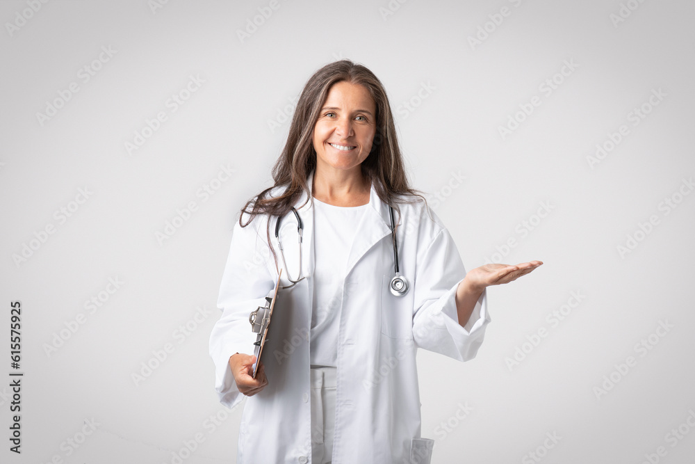 Excited senior woman doctor holding clipboard and showing something on empty palm, advertising poduct or clinic Excited senior woman doctor holding clipboard and showing something on empty palm, advertising poduct or clinic