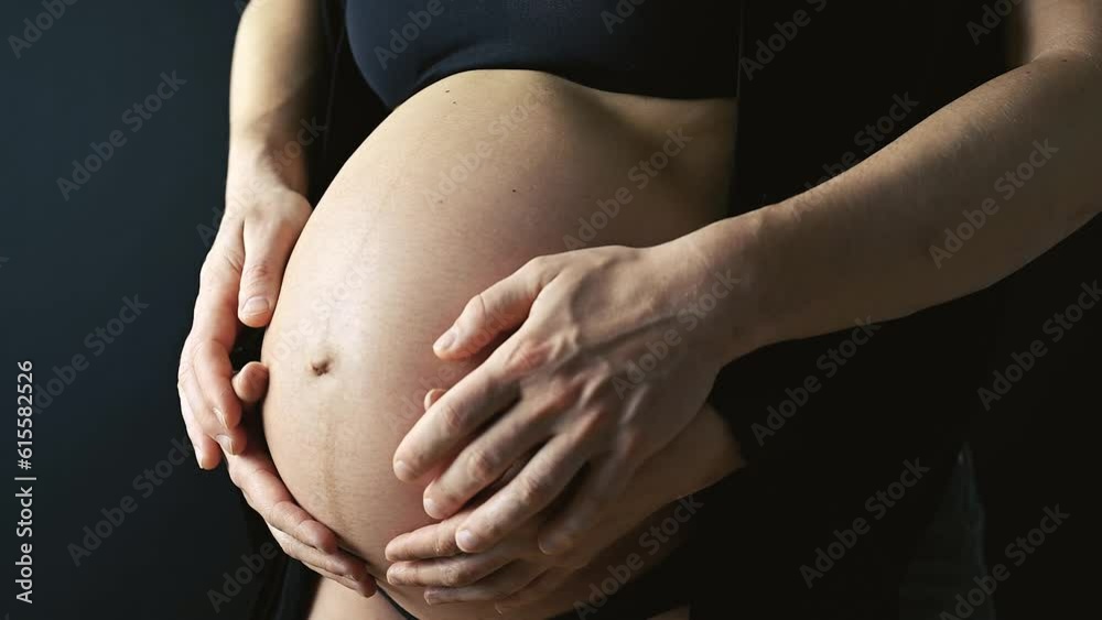 Closeup of naked round belly of pregnant mother in underwear held and carresed by mother and joined by fathers hands. Last month of pregnancy - 36th week. Side angle view. Black background. Closeup of naked round belly of pregnant mother in underwear held and carresed by mother and joined by fathers hands. Last month of pregnancy - 36th week. Side angle view. Black background.