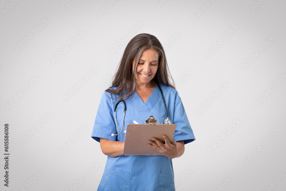 Happy senior female doc reviewing patient's medical history and taking notes to clipboard, light grey background - obrazy, fototapety, plakaty 