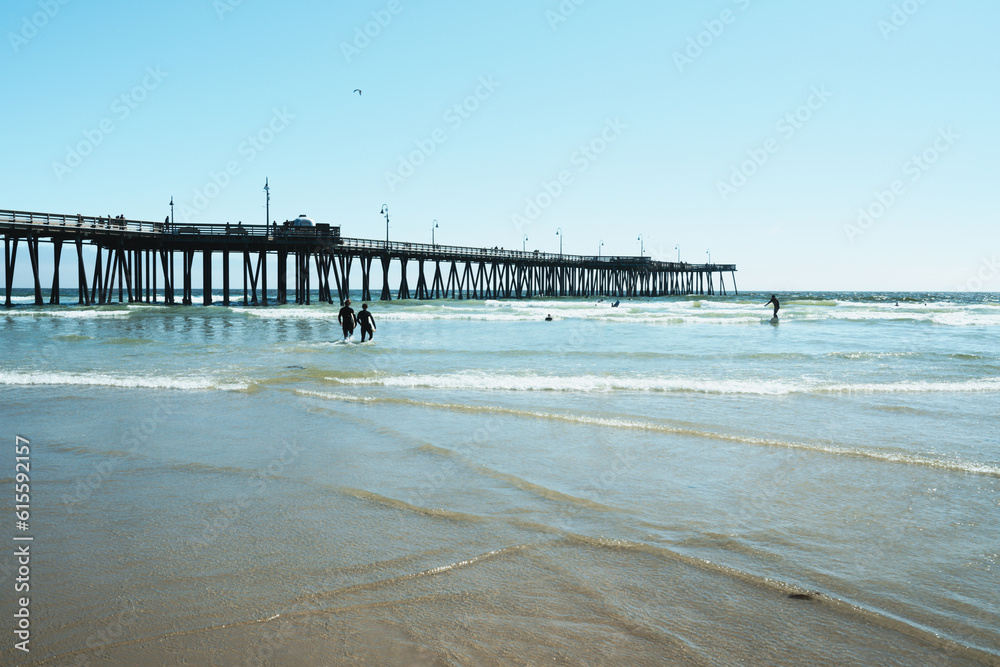 A long wooden pier, quiet ocean, silhouette of surfers on a bright sunny day