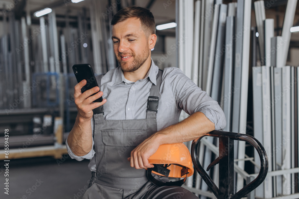 Portrait of a happy worker in a orange helmet and overalls holding a hydraulic truck and talking on the phone against a background of a factory and aluminum frames. Portrait of a happy worker in a orange helmet and overalls holding a hydraulic truck and talking on the phone against a background of a factory and aluminum frames.