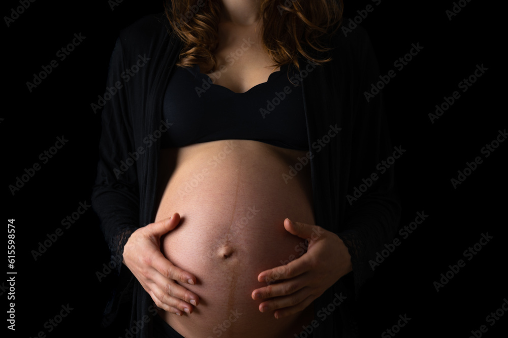 Atmospheric shot of unrecognizable pregnant woman in underwear gently holding her naked round belly. Last month of pregnancy - 36th week. Cropped, front view. Black background. Atmospheric shot of unrecognizable pregnant woman in underwear gently holding her naked round belly. Last month of pregnancy - 36th week. Cropped, front view. Black background.