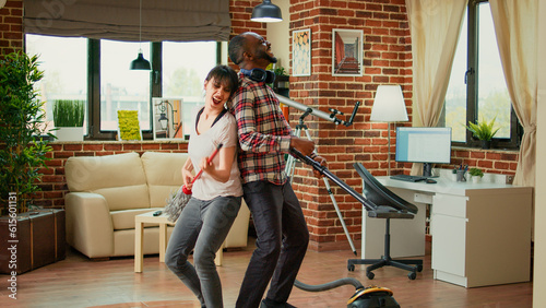 Cheerful people singing and cleaning apartment floors, using vacuum cleaner in living room. Young smiling couple dancing and listening to music, having fun with spring cleaning.