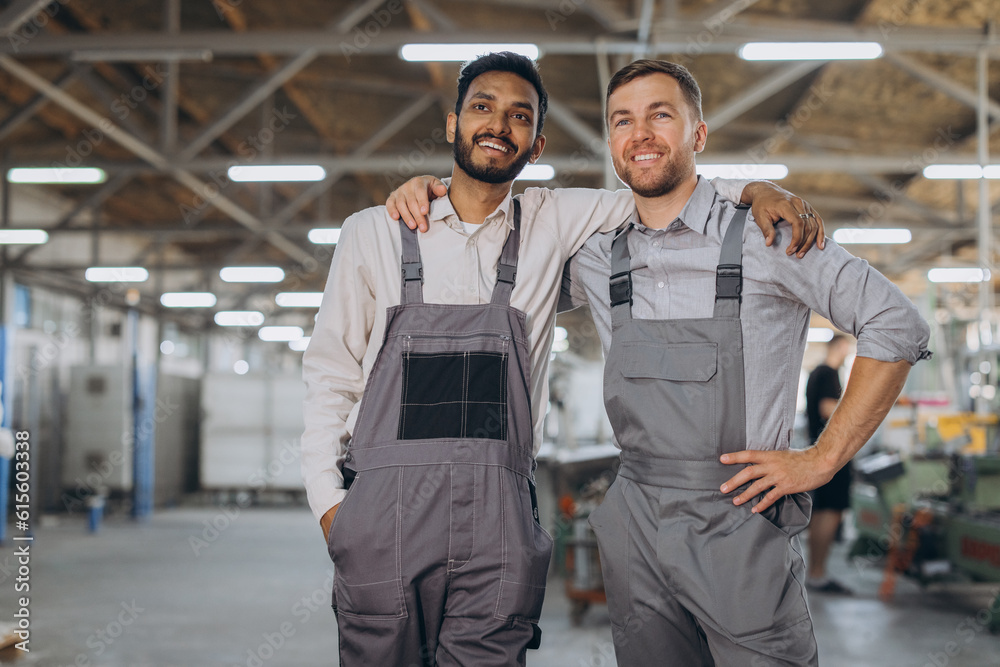 Two happy international workers in overalls hug each other against a factory background