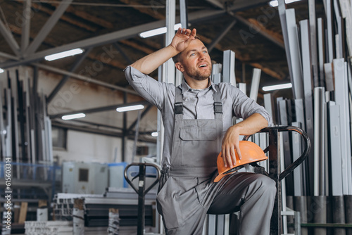 Portrait of happy worker in orange hard hat and overalls holding hydraulic truck and going on lunch break wiping sweat from forehead against factory background.