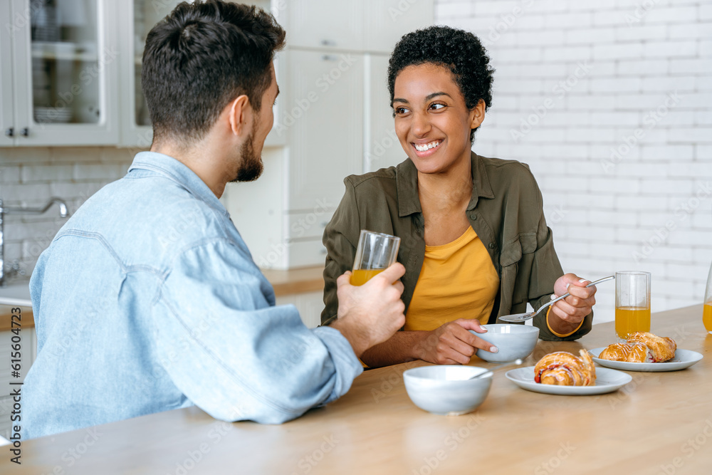 Weekend morning. Happy young family couple, caucasian man and african american woman, having breakfast at cozy home kitchen with healthy porridge, happy to spend time together, communicating, smile Weekend morning. Happy young family couple, caucasian man and african american woman, having breakfast at cozy home kitchen with healthy porridge, happy to spend time together, communicating, smile