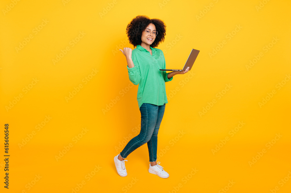 Full length photo of a stylish lovely african american or brazilian curly woman holding laptop and pointing finger back at the space for announcement, smiling, standing on isolated yellow background Full length photo of a stylish lovely african american or brazilian curly woman holding laptop and pointing finger back at the space for announcement, smiling, standing on isolated yellow background