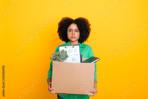 Unemployed. Dismissal from work. Upset unhappy brazilian or african american woman, fired from her job, holding box of office supplies, looking at camera sadly, standing on isolated yellow background