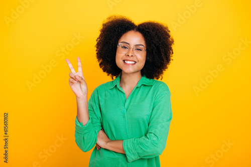 Beautiful positive curly young woman of brazilian or african american ethnicity, wearing green shirt, showing peace gesture with fingers, looks at camera, smiling, stand on isolated yellow background