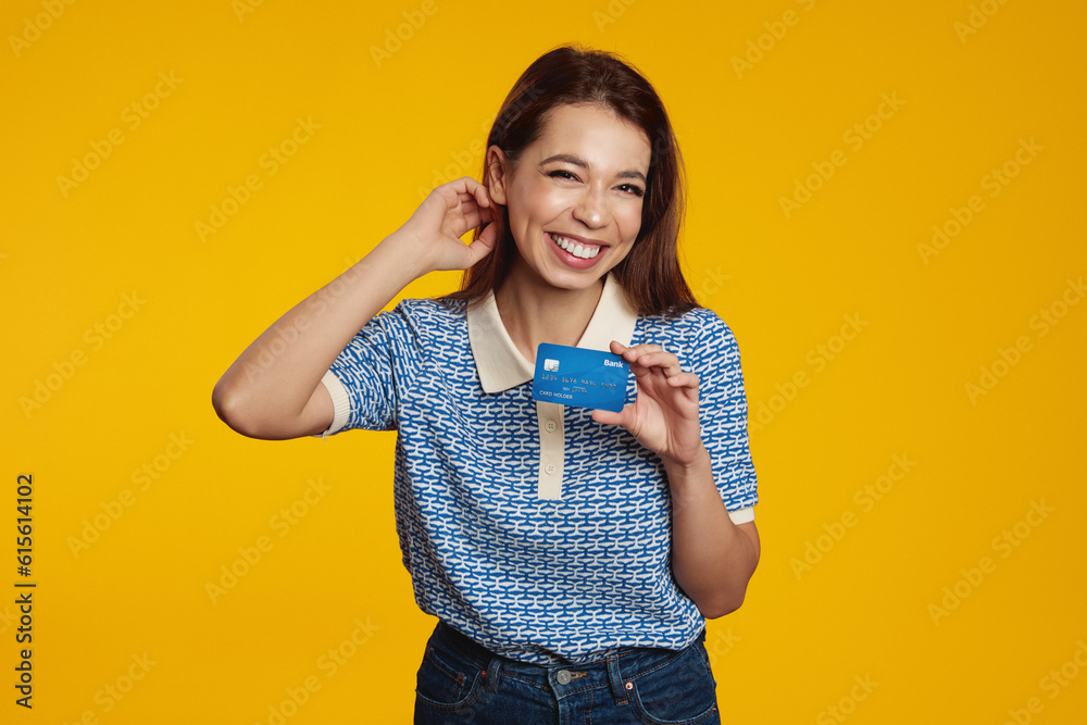 Smiling pretty brunette girl holding credit card while smiling and looking at camera over yellow background, wearing blue shirt Smiling pretty brunette girl holding credit card while smiling and looking at camera over yellow background, wearing blue shirt