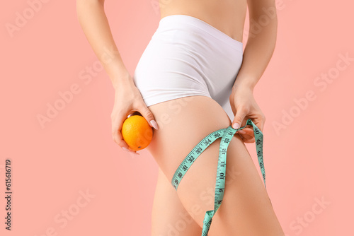 Young woman with orange measuring her thigh on pink background, closeup