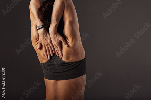 Female sporty muscular with ponytail doing stretching workout of the shoulders, blades in sport bra, holding the hands on the shoulders on grey background with empty copy space. Back view.