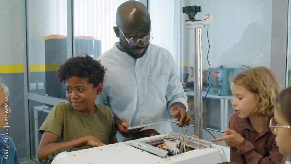Medium arc shot of African American male technology teacher showing wires and circuitry of delivery robot to group of multiethnic young boys and girls during lesson at afterschool robotics club Medium arc shot of African American male technology teacher showing wires and circuitry of delivery robot to group of multiethnic young boys and girls during lesson at afterschool robotics club