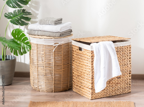 Two natural wicker laundry baskets filled with white towels, set on a wooden floor in a cozy room decorated with potted plants