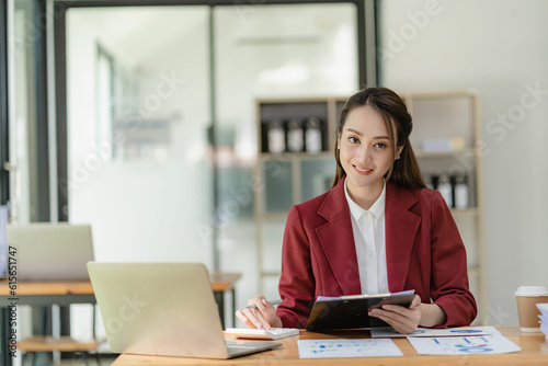 Asian businesswoman in suit sitting on desk in office, with computer document graph for bookkeeping in workplace to calculate annual profit by function, business concept