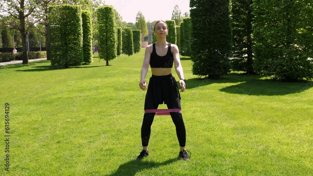 A young active woman in black sportswear with an elastic band on her legs trains in the park on the grass in summer. A woman squats on the lawn in a modern park. A young active woman in black sportswear with an elastic band on her legs trains in the park on the grass in summer. A woman squats on the lawn in a modern park.