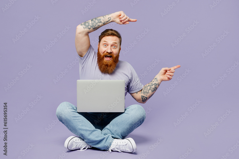 Full body young redhead bearded man in violet t-shirt casual clothes sitting hold use work on laptop pc computer point aside isolated on plain pastel light purple background studio Lifestyle concept - obrazy, fototapety, plakaty 