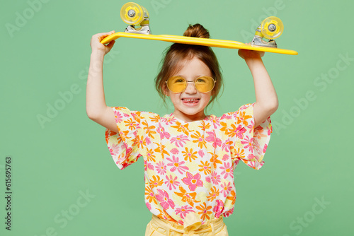 Little child kid girl 6-7 years old wears casual clothes sunglasses have fun hod in hand skateboard look aside on area isolated on plain green background. Mother's Day love family lifestyle concept. photo
