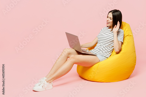 Full body young IT woman wear casual clothes t-shirt sit in bag chair hold use work on laptop pc computer waving hand isolated on plain pastel light pink background studio portrait. Lifestyle concept
