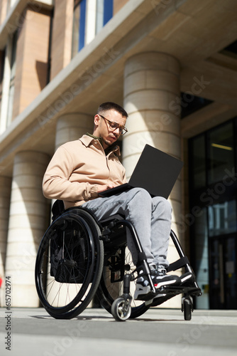 Vertical image of young man sitting in wheelchair and using laptop in the city © AnnaStills