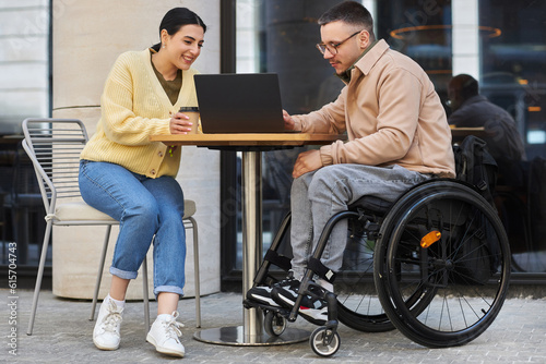 Young man with disability having business meeting with woman, they sitting in outdoor cafe and discussing online presentation on laptop