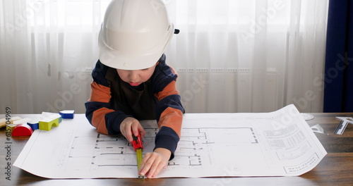 Preschooler in hardhat measures dimensions with tape measure on apartment drawing. Boy imitates role of engineer taking measurements with interest using tool on table