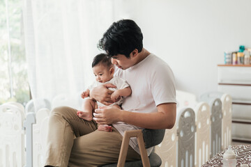 Happiness of fatherhood. Young Asian dad with adorable baby on his hands sitting near window at home. Loving father spending time with infant child. Fathers day concept.