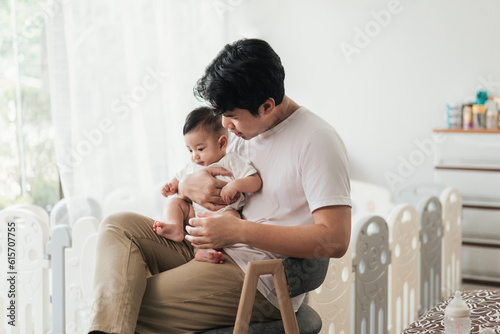 Happiness of fatherhood. Young Asian dad with adorable baby on his hands sitting near window at home. Loving father spending time with infant child. Fathers day concept.
