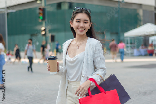 A beautiful and young asian female office employee smiles while walking around the city district. Holding a cup of coffee and some shopping bags.