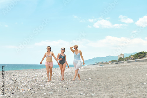Three women friends walking on the beach and laughing on a summer day. Cheerful females enjoying together summer vacation near the sea.