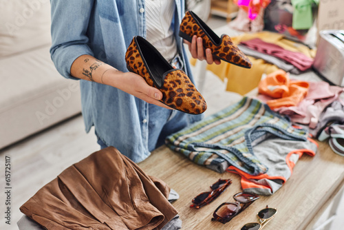 ethical consumption, exchange, cropped view of young tattooed woman holding stylish animal-print shoes near sunglasses and garments on table, sustainable living and mindful consumerism concept photo