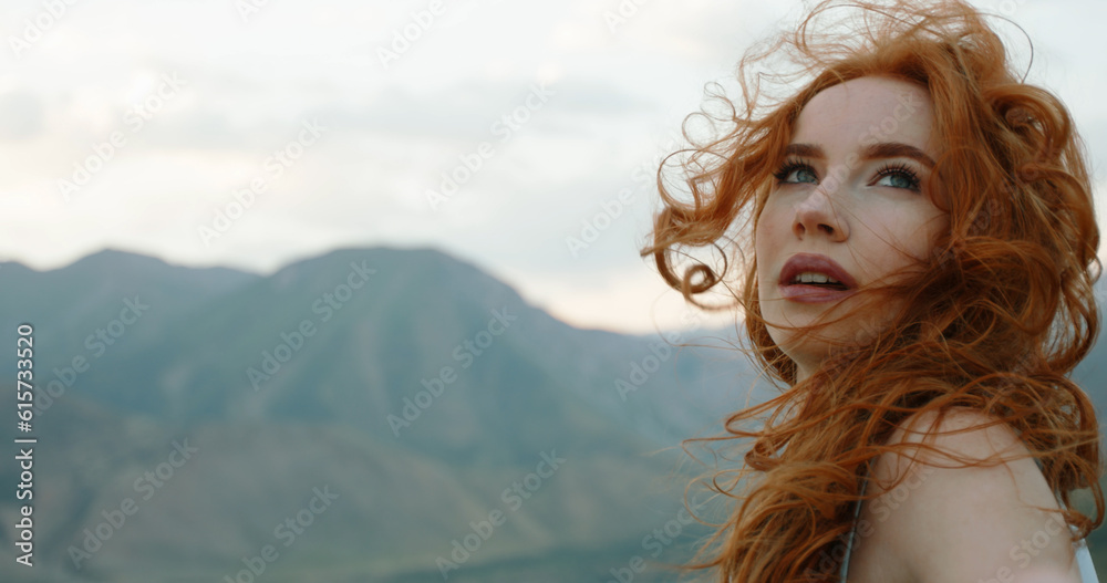 Beautiful female model on top of a mountain posing for photoshoot, reaching her hand out to camera while wind blows her dress and red hair - freedom, tranquility. Copy Space