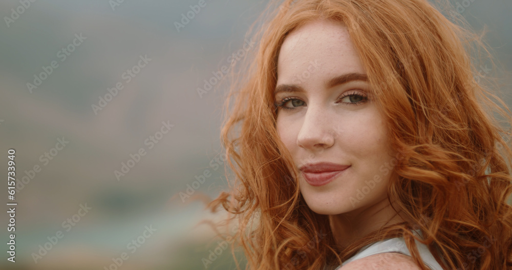Close up shot of face of beautiful young caucasian girl with red hair and freckles looking at camera and positively smiling. Cute woman on portrait in nature. Copy Space