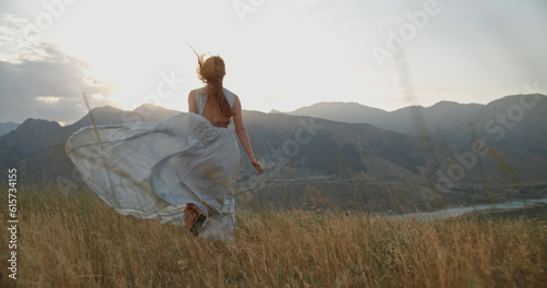 Young beautiful girl with red hair wearing white dress running on top of a mountain facing wind blowing her hair and dress and smiling - freedom, adventure, harmony 