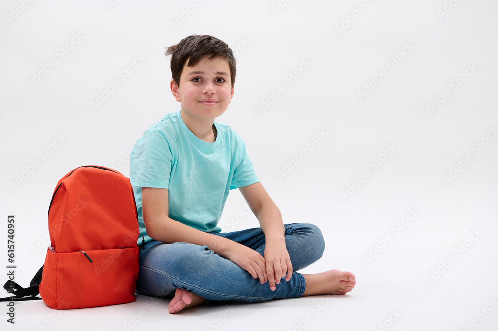 Caucasian confident smart teen boy in blue t-shirt and jeans with orange rucksack, smiling looking at camera, sitting barefoot over isolated white studio background. Portrait of a schoolboy. Education Caucasian confident smart teen boy in blue t-shirt and jeans with orange rucksack, smiling looking at camera, sitting barefoot over isolated white studio background. Portrait of a schoolboy. Education