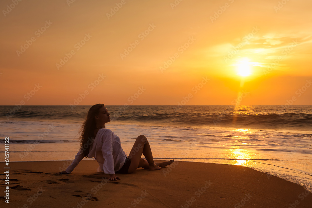 Silhouette of slim woman sitting on tropical beach at ocean sunset, rear side view. Slender lady in beachwear relaxing enjoying tropic seaside, looks away. Travel vacation concept. Copy ad text space