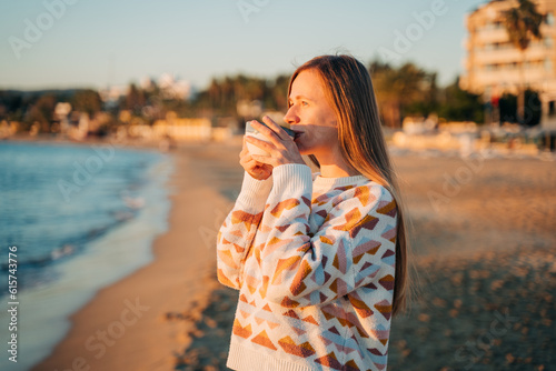 Young beautiful girl in cozy sweater drinking coffee on a winter seaside shore while taking sunbathe in a mild sunset. Cute attractive woman enjoying her cup of tea while watching autumn ocean waves.