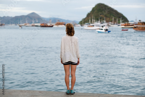Girl traveler of European appearance, enjoys life, looking at the sea bay with yachts, rear view.