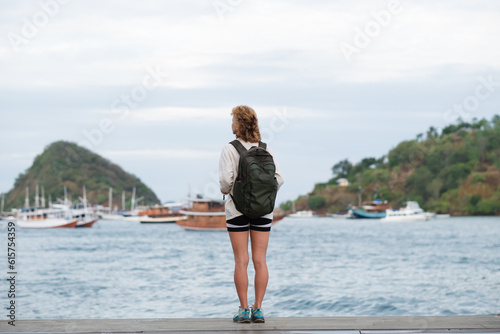 A girl traveler of European appearance, with a backpack, looks at the sea bay with yachts, rear view.